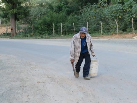 Texto alternativo: Pescador de edad avanzada camina rumbo a Estero Balsa, por una carretera de Manzanillo cargando un cubo y una herramienta de pesca, símbolo de las dificultades actuales para acceder a las zonas tradicionales de pesca.